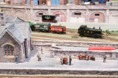 Passengers waiting on the station platform. The station entrance can be seen on the end of the building, with steps from the lower road level.